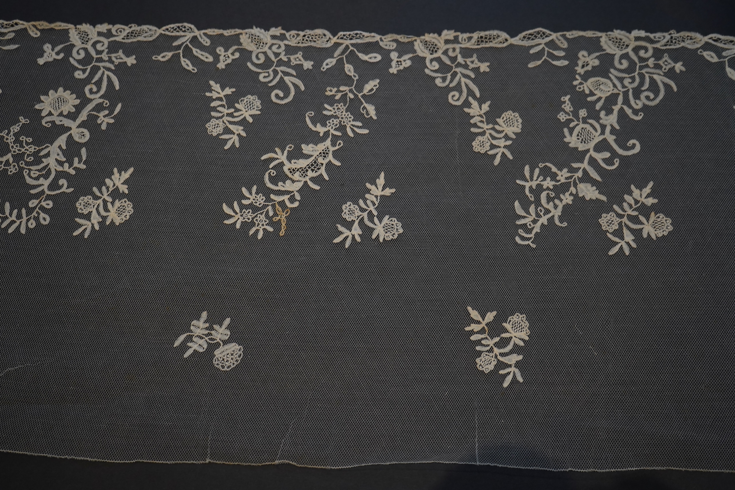 A 19th century five yard flounce of Brussels bobbin appliqué lace applied onto fine machine net, worked in a trailing floral design with an ornate border and sprig motifs, 457.5 cm long x 36cm deep. Condition - possibly